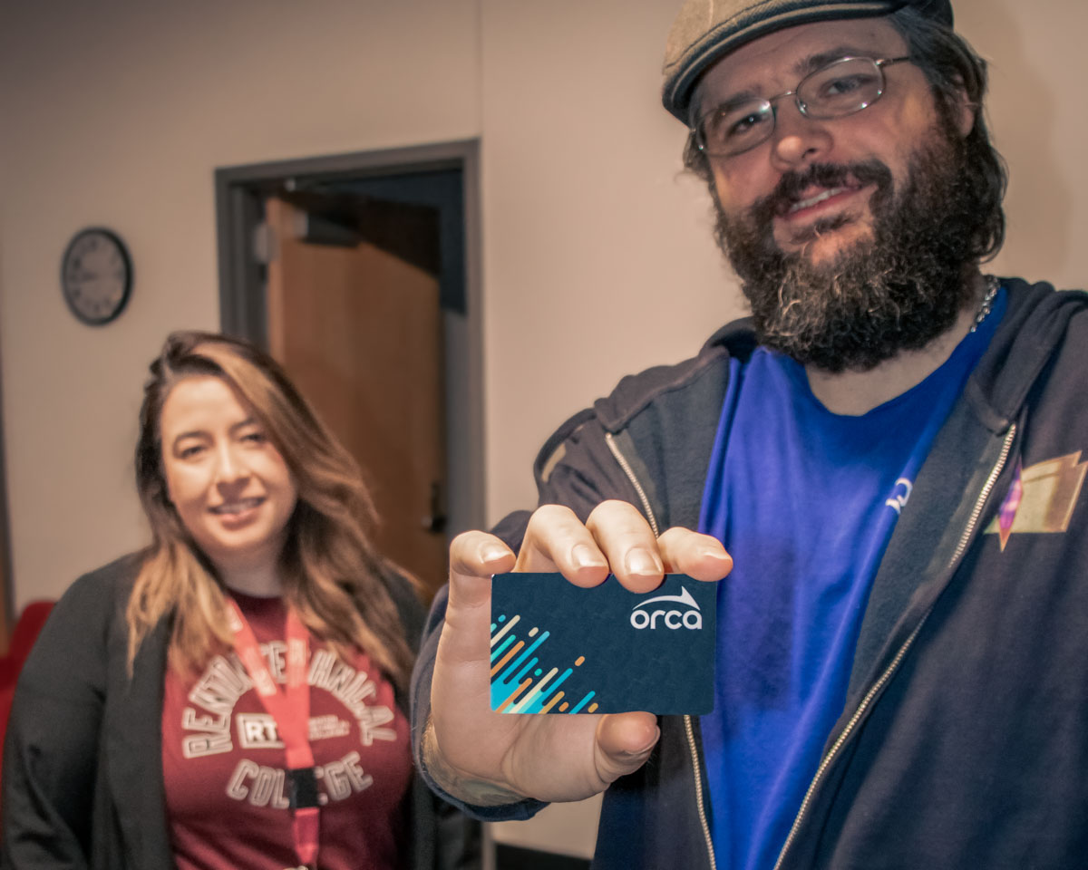Student holding an ORCA transit card toward the camera, with another person standing in the background smiling