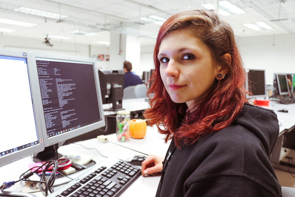 Anna looking at the camera, a computer monitor in front of her with lines of codes 